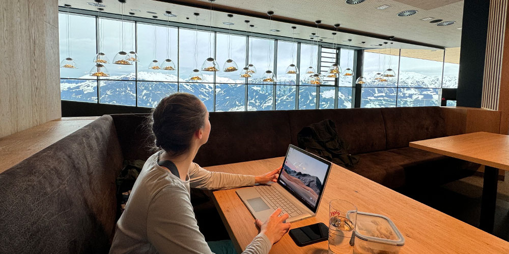Elena sitzt am Tisch mit ihrem Laptop und hat dabei einen Ausblick auf die Berge | Ski-Office | Kraus & Partner - Transformation Experts